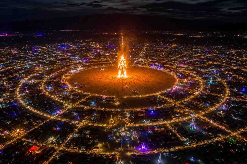 Aerial view of Black Rock City at night during Burning Man 2026 festival with the Man burning in center
