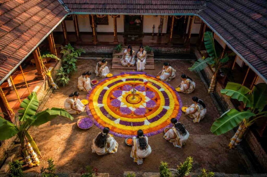 Women in kasavu sarees standing around a colorful onam pookalam in a Kerala courtyard