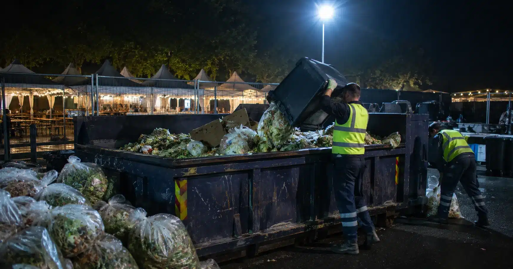 Workers loading bagged food waste into a skip behind a festival food court at night