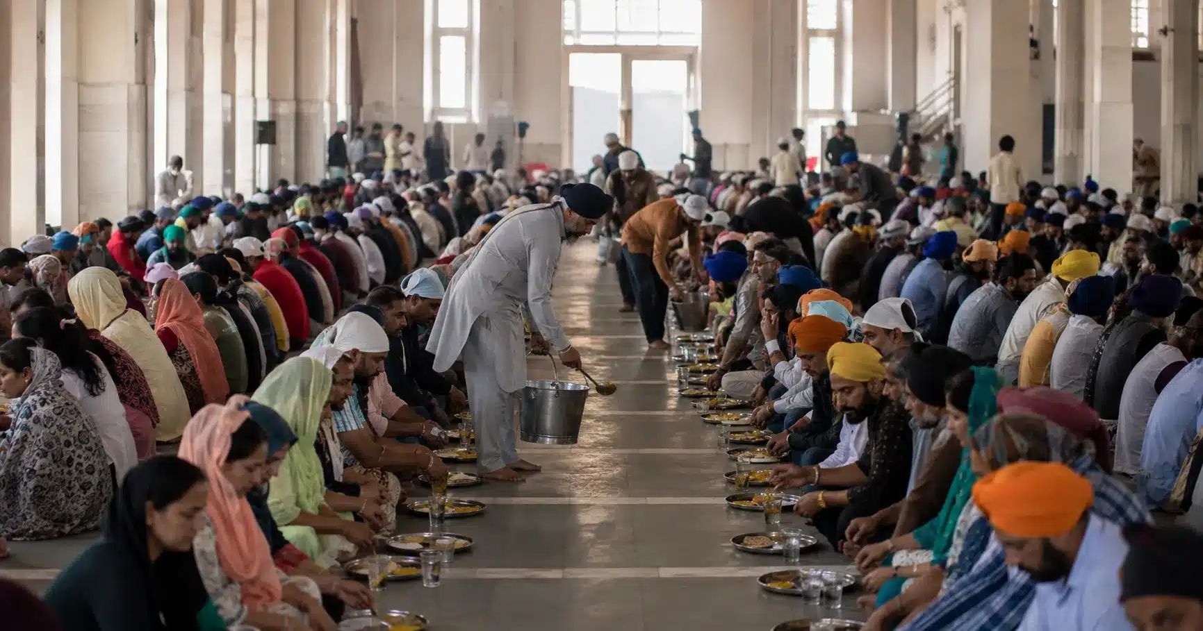Volunteers serving food to rows of people seated on the floor in a langar community kitchen hall