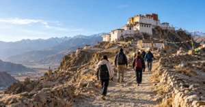 Visitors in layered clothing and daypacks walking a stone path toward a whitewashed monastery on a dry mountain ridge